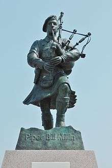 Photograph of a life-sized bronze statue with a greenish verdigris patina depicting Millin marching in his kilt and playing his bagpipes
