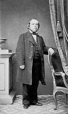 Photo of Louis Plaidy wearing a long coat, standing by a chair.
