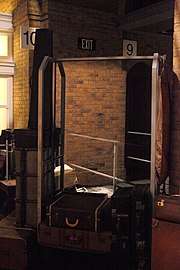 Photograph of a hotel-style metal-framed luggage trolley surrounded by old-style suitcases. In the background is what appears to be a solid brick wall. with platform number sign "10" on the left, and "9" on the right.  The trolley contains a human-sized semi-transparent mirror that combines the foreground and background.