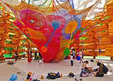 A weblike structure in many bright colors with several pendulous appendages hanging from it, itself hanging from the textile roof of an open wooden structure. There are children playing with the pendulous appendages and seated around the side, where many backpacks and shoes have also been left