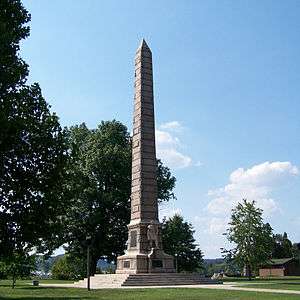 A stone obelisk monument against a blue sky and trees, with the statue of a pioneer in the front.