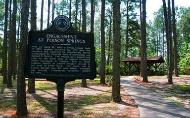 Historic marker reading "Engagement at Poison Springs" in foreground of a forested area with rustic wooden pavilion in the background.
