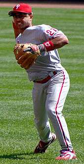 A man wearing a gray baseball uniform with "Phillies" across the chest in red script and a red baseball cap throws a baseball with his right hand.