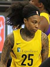 Waist high portrait of young woman with tatoos on her arms wearing yellow basketball uniform with lot of dark hair swept up into a high ponytail