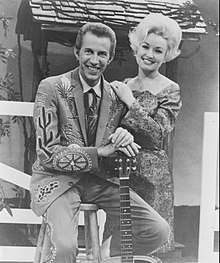 A man in an elaborately-patterned suit, sitting on a stool and leaning on the neck of a guitar.  A blonde woman stands to his left, resting her hands on his shoulder.