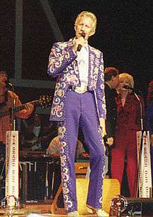 A blond-haired man wearing a blue suit with elaborate patterns on the jacket and the sides of the pants, singing into a microphone