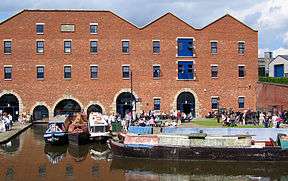 A brick-built warehouse with a canal in the foreground. There are barges on the canal and people sitting outside the warehouse.