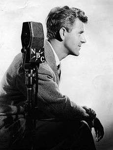 Side view of a white male wearing a suit jacket. A microphone marked with the letters NBC is placed in front of the subject.