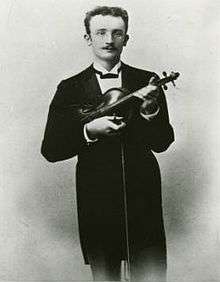 A thirtysomething man in a tuxedo and glasses clutches a violin and gazes at the camera.
