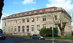 Photograph of the Astoria post office, a broad, institutional, two-story building in concrete or stone with a low-profile, red roof