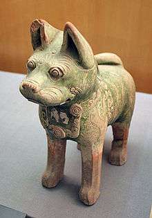 A green-glazed ceramic statuette of a dog with pointy ears and curly tail, standing upright on all fours with eyes open