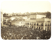 An old photograph showing a crowded square in front of a large, white, multi-storied building