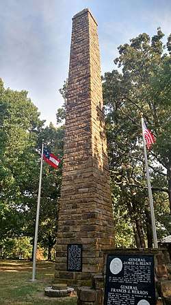 Tall field stone column memorial flanked by United States and Confederate States of America flags surrounded by tall, leafy green trees.