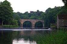 A stone two-arched bridge across a river, viewed along the river, both ends hidden by trees. A weir is in front of the bridge, at the right end of which is a two-storey building.