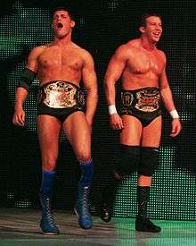 Two dark-haired men are both wearing black wrestling tights. One wears knee-high blue wrestling boots with blue knee-pads and a black elbow pad on his right arm, and the other wears black knee-high wrestling boots with black knee-pads, and white tape around his wrists. The two are wearing professional wrestling championship belts around their waists.