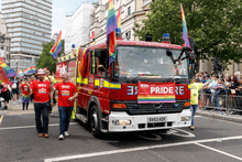 Two firefighters in red "LFB PRIDE" tshirts walk in a parade alongside a fire engine. The fire engine is decorated with at least five rainbow flags.