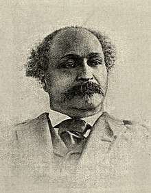 Bust-length photographic portrait of a black man with a mustache and slight tuft of facial hair at his lower lip, wearing a gray suit and dark silken necktie against an illuminated white background. His head is bald at the top with locks of greyed curly hair sticking out on both sides of his head behind his temples