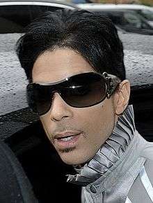 A color photograph of Prince, who is peering into a nearby camera.