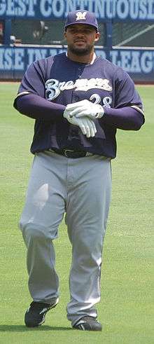 A man wearing a navy blue Brewers jersey, gray pants, and a navy blue cap adjusts his white batting gloves.