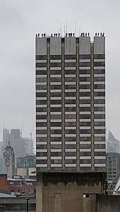 A photograph of the Project84 installation in London. A tall tower block - at the apex, statues of men stand disturbingly close to the edge.