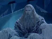 A man sits frozen in an icy throne room, he is covered in ice as icicles hang from his clothes and long beard.