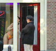 A photograph depicting a woman wearing an orange bikini leaning out of an open doorway in front of a man wearing a black jacket