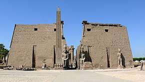 Pair of trapezoidal stone towers flanking a passage, beyond which a row of columns is visible. In front of the towers are several large statues and an obelisk.