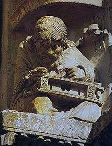 Medieval carving of a man with long hair and a long beard hunched over a musical instrument he is working on