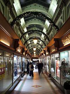 An ornate shopping arcade, lit up by ground floor shop windows, shop signs and fairy lights from above, in the tall ceiling space. The floor is surfaced with white tiles, with linear patterns along the edges of the walkway and diamonds interspersed along the centre.