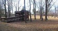 An open wooden structure with a ramp leading up to it, on the edge of a large open field with large leafless trees around