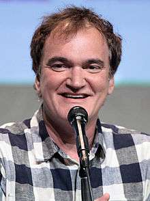 A smiling Quentin Tarantino dressed in a casual shirt speaking into a microphone