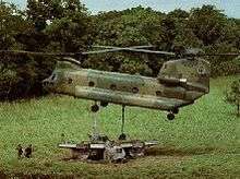 Colour photo of a twin-rotored helicopter flying just above the wreckage of an aircraft