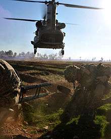 colour photograph of two marines in a field ducking as a low-flying helicopter fluies overhead.