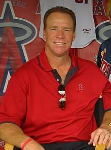A man in a red shirt smiles with a pair of sunglasses folded in the front of his shirt.