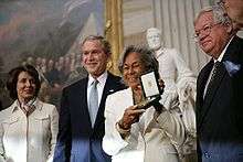 A dark-skinned woman holds a medal in a green case. She is standing in a group of four people and has a wide smile. The woman has wavy gray hair and is dressed in a white suit.