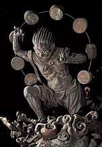 Raijin. Three-quarter view of a statue. His left leg is bend as if climbing stairs and he is leaning forward. His hair is standing up and his right hand is raised above his head. A thin halo around his head is decorated with thick circular disks. Black and white photograph.