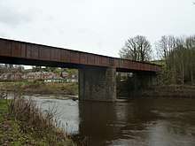 A rusty plate girder bridge crosses the river, supported by a single stone pier
