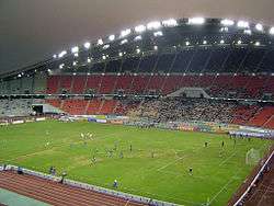 A stadium with green field for football, an athletic track, sitting area and floodlights; a football match is in progress