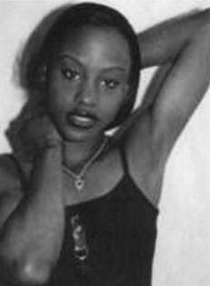 A black and white photo of a dark-skinned woman wearing a dark halter top, seen from the bust up, with one hand at her neck and the other behind her head