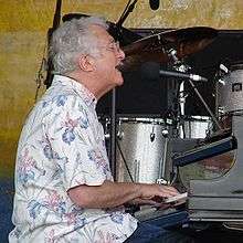 Side profile of an elderly grey-haired man seated to a grand piano, which he is playing while singing into a microphone; he is wearing a floral shirt.