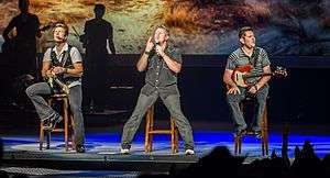 Three men performing music on a stage.  One is singing and the others are playing guitars.  All three are sitting on stools.