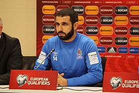 A bearded man in a blue top sits behind a desk at a press conference