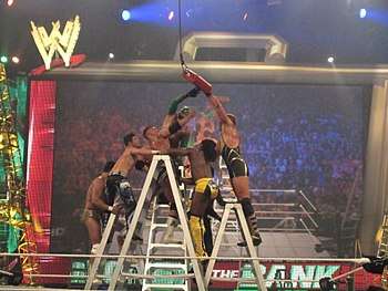Seven professional wrestlers battle on top of four ladders in the ring, reaching for a red briefcase hanging above them.