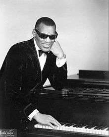 A dark-skinned man wearing dark glasses and a tuxedo, leaning on a piano