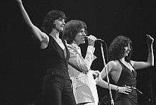 Three people sing onstage next to each other. The man in the middle wears white clothes, and on both sides stands a woman wearing black.