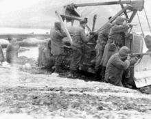 A group of eight or nine men, some standing some sitting, most taking cover behind a bulldozer and with rifles in firing position, in a barren landscape with both water and mountains in the distance