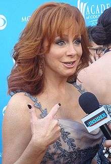 A woman with long red hair, wearing a grey dress, talking into a microphone
