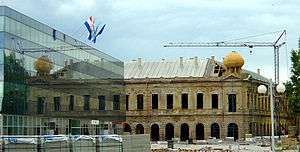 A large damaged building under restoration with a modern glass building visible in the foreground, flying various flags. A construction crane and building materials are also visible.