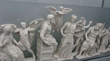 Une dizaine de statues blanches alignées : au centre un homme barbu, face à lui une femme en armes ; entre eux une figure féminine en vol ; divers autres personnages