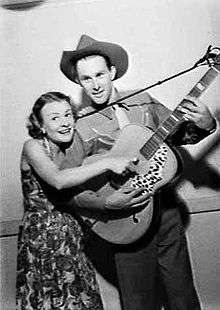 A three-quarter black-and-white image of a couple. The woman at left is wearing a floral dress and has her right hand placed on the string of the guitar. It is being held by the twenty-five year-old man who wears a cowboy shirt and a hat. His left hand is on the fret board and his right is below the woman's hand.